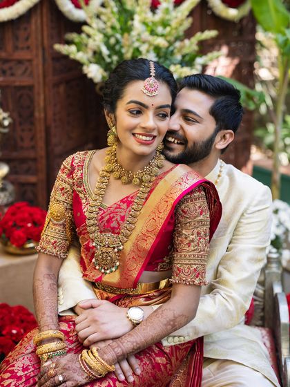 A happy couple posing for a photo. Her red Kanchipuram silk saree with a heavily worked blouse is a classic bridal choice, complemented by his elegant cream kurta.