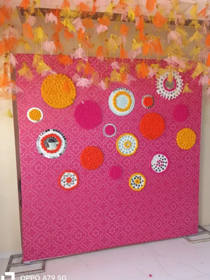 A creative and colorful photo wall for a mehendi or sangeet. The pink patterned backdrop is decorated with circular patterns made from marigold flowers and mirrors.