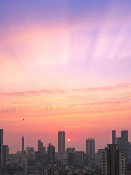 Birds fly across a stunning purple and pink sunset sky over the Mumbai skyline, with sun rays breaking through the clouds.