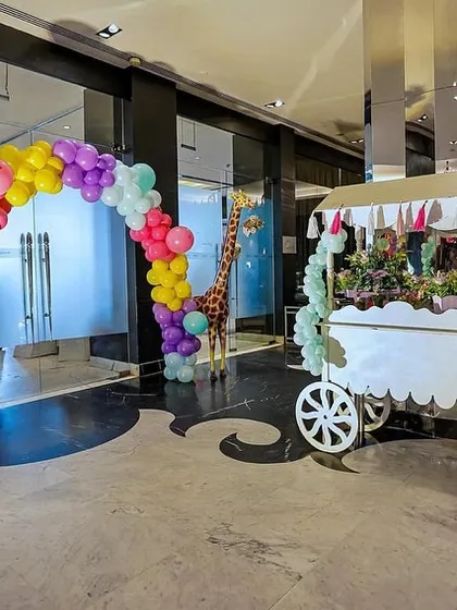 A vibrant entrance to a carnival-themed party. A colorful balloon arch frames the walkway, leading guests past life-sized giraffes and a directional signpost cart, setting a festive tone from the start.