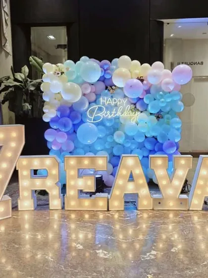 A vibrant and colorful setup for Reava's 7th birthday. The wall of pastel blue and purple balloons paired with large, lit-up marquee letters of her name creates a fun and personalized photo backdrop.