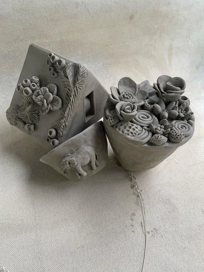 A collection of intricate, unfired hand-built sculptures. This image showcases a house with detailed floral work, a succulent planter, and a piece with an elephant motif.