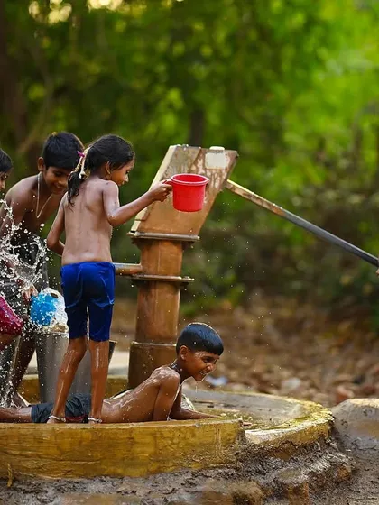 The scene captures multiple stories at once: one boy pumps water, others splash, and one lies down to cool off. It’s a complete narrative of a summer afternoon at the well.