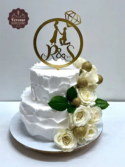This elegant two-tier engagement cake features a textured white whipped cream frosting and a cascade of fresh white roses with glittery gold balls. A custom topper with the couple's initials adds a personal touch.