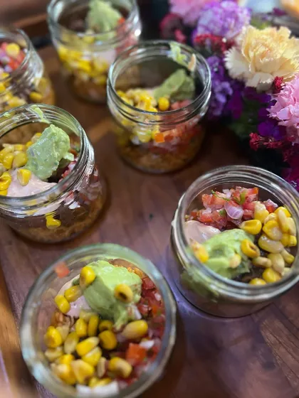 Our Mexican seven-layer dip served in individual jars. These are perfect for guests to grab and enjoy, featuring layers of beans, guacamole, salsa, and corn. It's a fun and interactive addition to our catering menu.