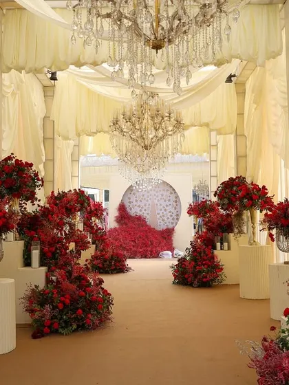 This entryway makes a bold and opulent statement. The path is flanked by towering arrangements of rich red florals, leading into a hall draped in ivory and illuminated by grand chandeliers, creating an immediate sense of luxury and drama.