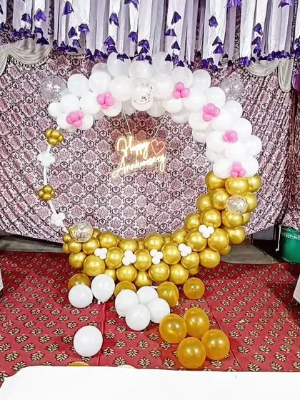 A circular anniversary backdrop made of gold and white balloons, with delicate pink accents. The 'Happy Anniversary' neon sign adds a modern, romantic touch.