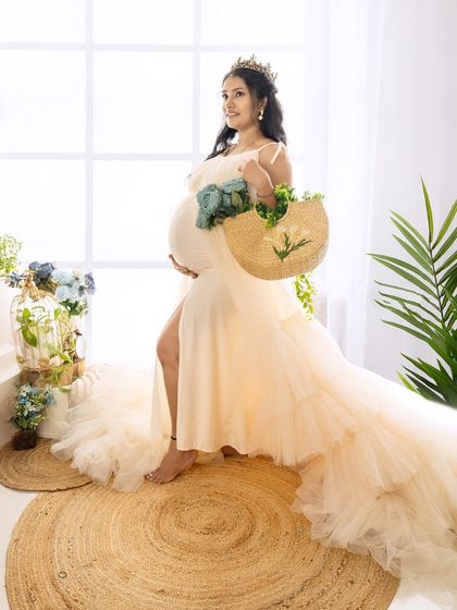 A full-length shot of a mom-to-be in a beautiful cream gown, wearing a crown and holding a stylish bag of flowers.