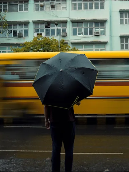 A person stands with an umbrella as a yellow bus blurs past. The strong color contrast and motion blur make this a dynamic and eye-catching street photograph.