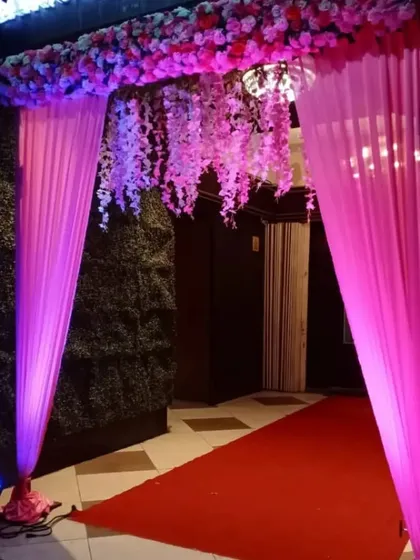 A classic entrance gate decoration for an indoor venue. We used bright pink drapes and an overhead arrangement of artificial flowers with hanging wisteria to create a welcoming and festive entryway.