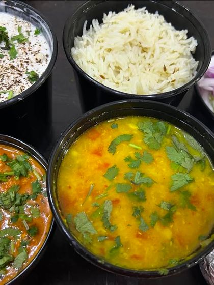 This is how I serve a complete meal for a family. It includes dal, paneer sabzi, jeera rice, fresh raita, salad, and rotis, all packed in separate containers.