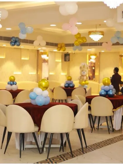 This image shows our hall set up for a birthday or baby shower celebration. The space is decorated with pastel blue, white, and gold balloons, with round tables covered in deep red cloths, creating a festive and welcoming environment.