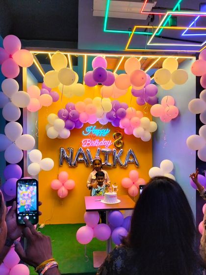 A candid moment from a birthday party in a cafe. The pastel balloon backdrop with the child's name creates a lovely setting for cake cutting.