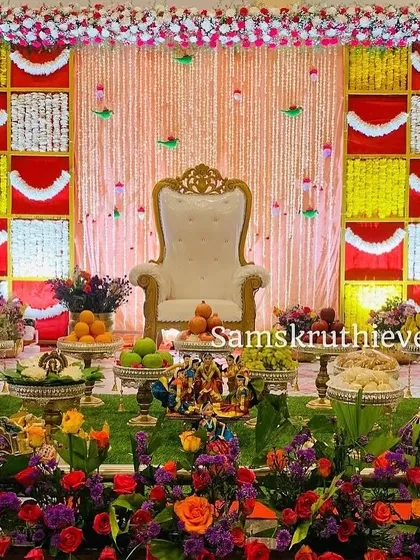 A full stage view of a baby shower with a red and yellow checkerboard backdrop. The floor is covered with a lush flower bed, and the offering table is filled with traditional items, creating a vibrant and complete look.
