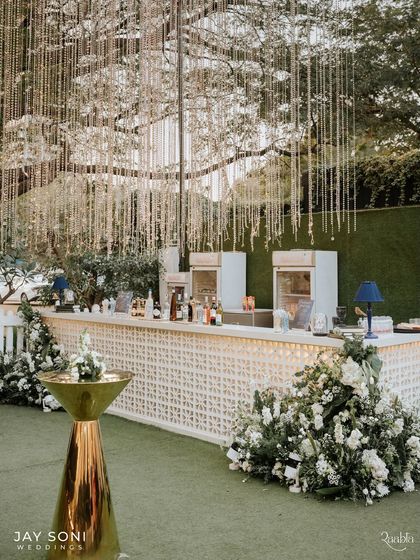 The bar area is an important part of the guest experience. I designed this one with a clean, white lattice front, a brass cocktail table, and lush floral arrangements, all under a canopy of hanging lights.