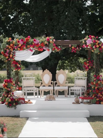 This stunning outdoor mandap combines rustic charm with vibrant tradition. The sturdy wooden frame is adorned with a cascade of bright red, pink, and orange flowers, creating a beautiful and sacred space for a wedding ceremony.
