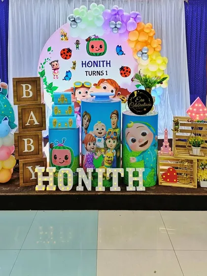 A full view of the Cocomelon first birthday setup. It includes themed plinths, wooden 'BABY' blocks, a character cutout, and a colorful balloon garland.