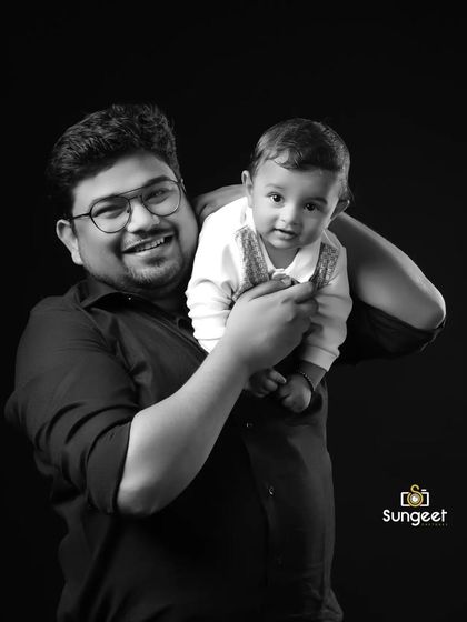 A classic black and white portrait of a father and his baby. The simple black background and focused lighting highlight the joyful interaction and the strong bond between them.