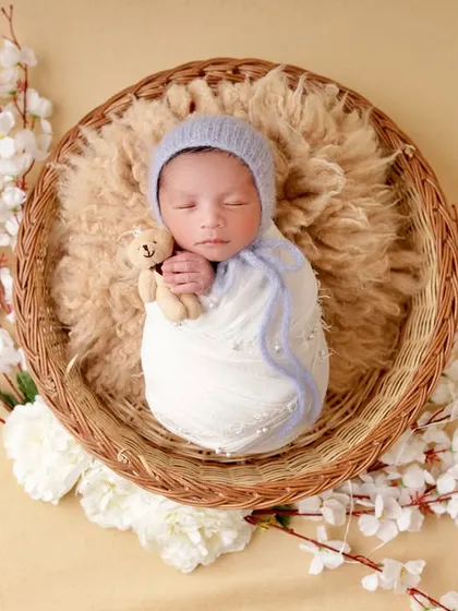 A baby boy in a soft blue bonnet cuddles a tiny teddy bear while sleeping in a wicker basket. The cherry blossom branches add a delicate, artistic touch to the scene.