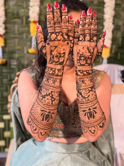 A playful pose from a bride showing off her full-arm mehendi. The design features bold lotus patterns and dense, traditional work.