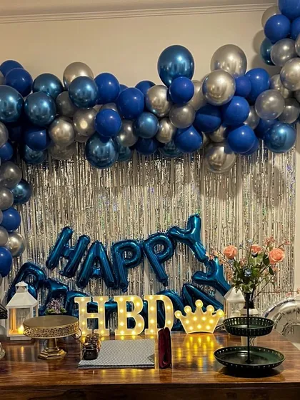 A classic blue and silver theme perfect for a stylish birthday celebration at home. The setup includes a balloon arch against a silver fringe curtain and light-up 'HBD' letters.