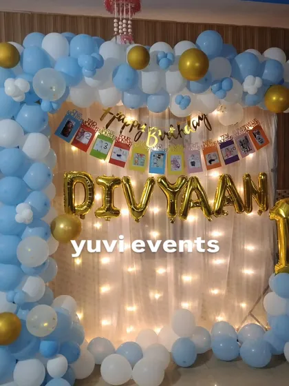 A simple and sweet first birthday decoration at home, with a blue and white balloon frame, fairy lights, and a personalized photo banner.