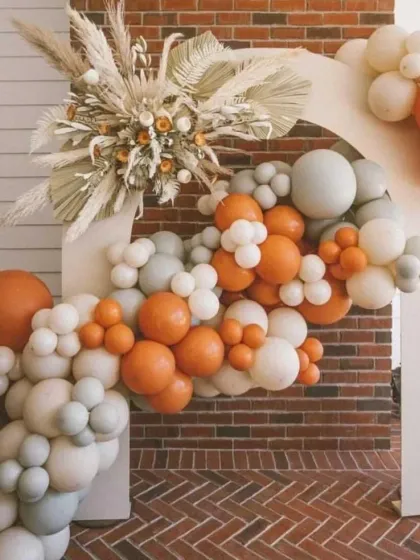 A modern and stylish baby shower backdrop featuring a boho-inspired balloon arch in burnt orange and sage green, complemented by dried pampas grass.