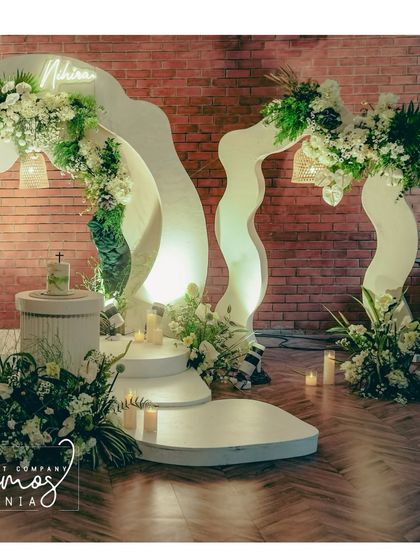 A modern and artistic baptism stage with white, wavy structures and lush green and white floral arrangements. The personalized neon sign adds a contemporary and heartfelt touch.