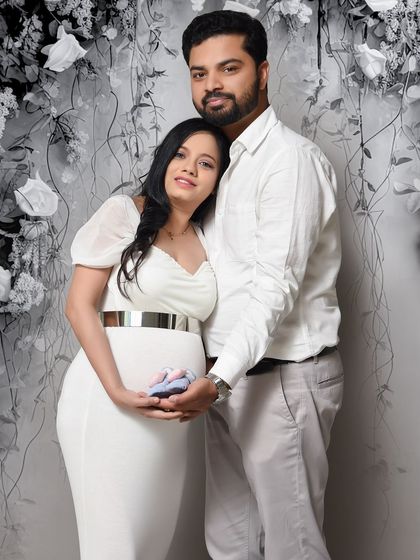 A classic couple's portrait against a grey floral backdrop. Dressed in white, they hold a tiny pair of baby shoes, symbolizing their journey into parenthood.