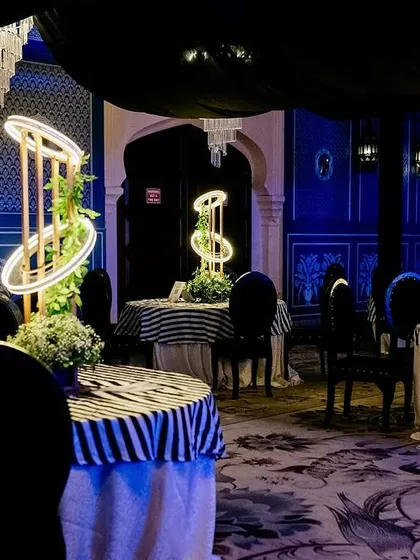 A chic Sangeet seating area with black and white striped tablecloths, modern illuminated centerpieces, and a backdrop of royal blue walls with traditional motifs.