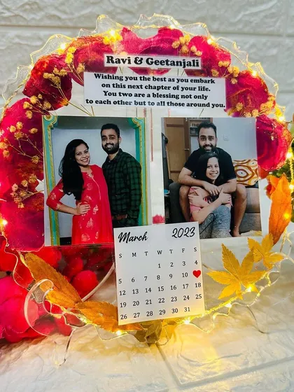 A round tabletop frame with preserved red rose petals, two photos, a custom message, and a calendar marking a special date. The frame is illuminated with fairy lights.