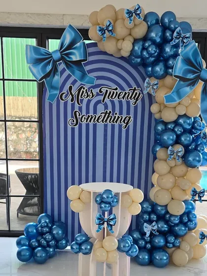 A chic and modern "Miss Twenty Something" birthday setup with a striped blue backdrop and a cream and blue balloon arch adorned with bows.