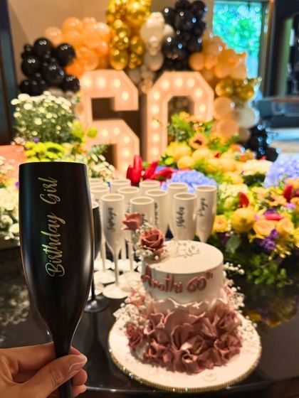A toast to a 60th birthday with a personalized "Birthday Girl" champagne flute. In the background, the dessert table is lavishly decorated with a custom cake, floral arrangements, and marquee numbers.