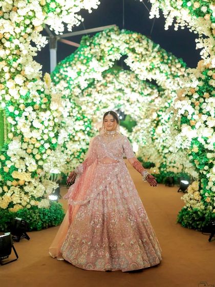 The bride standing under a magnificent floral arch. This full-length shot shows how the makeup complements her grand, pastel pink lehenga.
