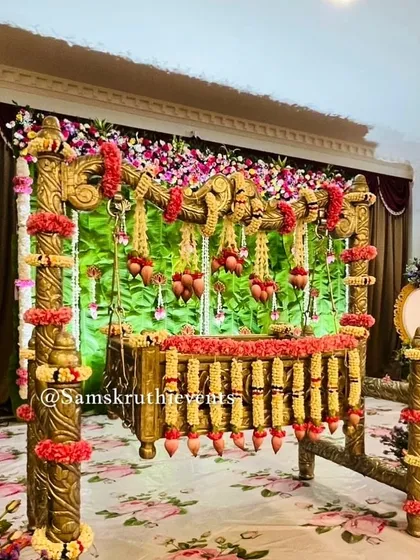 A traditional wooden cradle decorated with garlands of red and yellow flowers. The green backdrop and floral carpet create a vibrant and natural setting for the baby's naming ceremony.