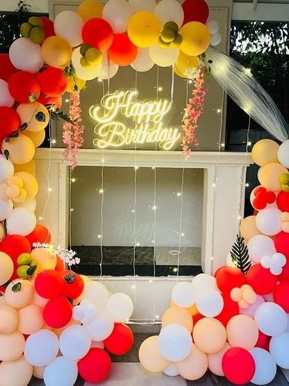 A warm and inviting balloon arch in red, orange, and white. The setup is framed by a white structure and accented with fairy lights and small floral details.