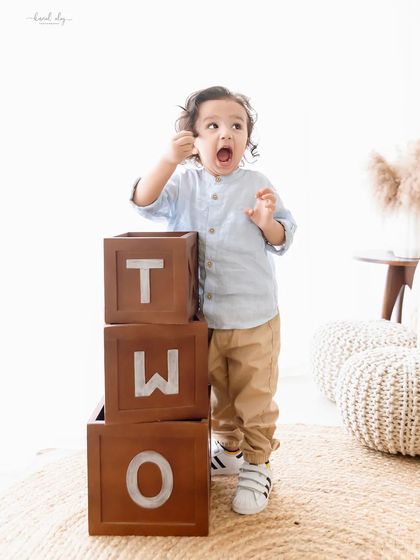 A toddler makes an excited face while standing next to "TWO" blocks, marking his second birthday.