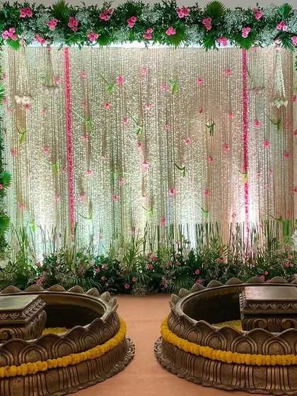 An indoor Haldi setup featuring a backdrop of delicate white and pink flower strings. The stage is flanked by floral arrangements on stands, with two large brass lotus-shaped urlis for the ceremony.