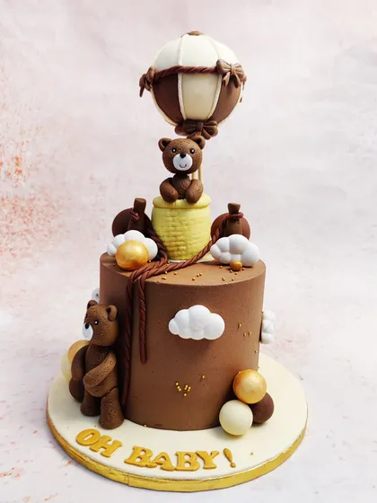 This Hot Air Balloon Theme Cake features two teddy bears, one flying high and another on the ground, creating a sweet "Oh Baby!" scene.