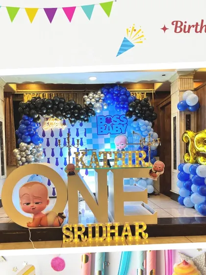 A fun and detailed Boss Baby themed first birthday setup. The stage includes a custom backdrop, large "ONE" lettering with the main character, and a blue, black, and silver balloon arch.