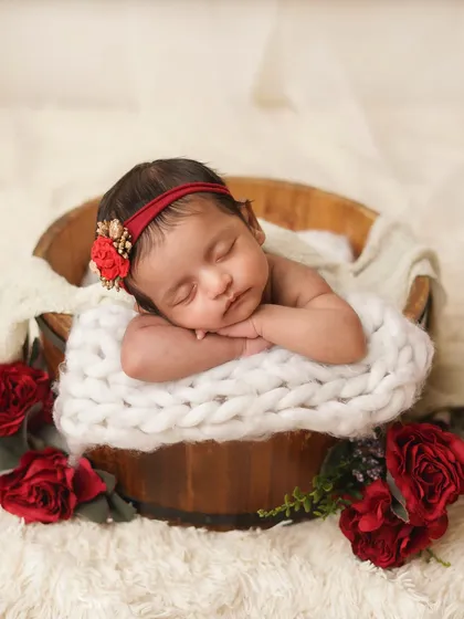 A newborn girl posed with her head on her hands in a bucket, framed by beautiful red roses. This is a classic and beloved pose in newborn photography.