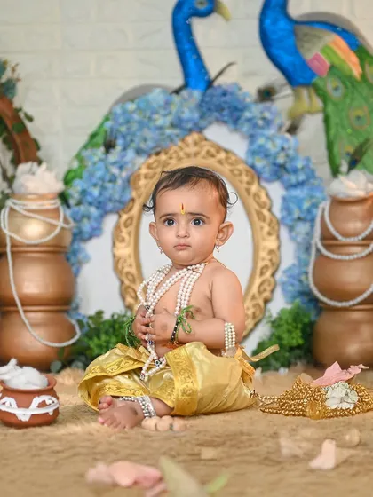 A classic Krishna pose with pots of butter and peacock decor. The baby's innocent expression is perfectly captured.