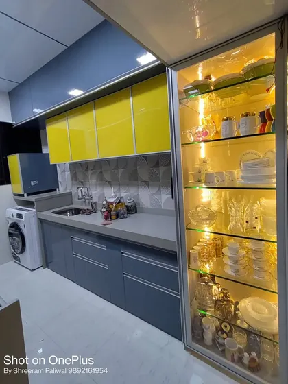 A vibrant kitchen design in Kandivali combining lemon yellow and grey high-gloss cabinets. The illuminated glass crockery unit on the right provides a stunning display for fine dinnerware.