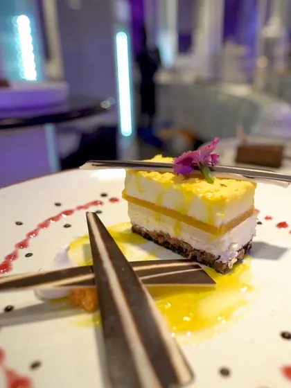 A slice of mango cheesecake, artfully deconstructed with chocolate shards and berry coulis. A tropical flavour presented with architectural precision.