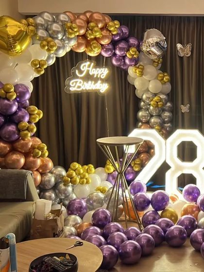 A stylish 28th birthday decoration in a living room. The setup features a circular balloon arch in purple, gold, and silver, a neon sign, and large '28' marquee numbers.
