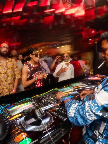 A long exposure shot capturing the movement and energy behind the decks. The blur of the crowd and lights reflects the fast-paced vibe of the night.