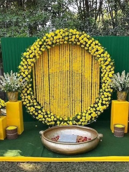 A classic Haldi backdrop for outdoor functions. This setup features a large circular frame filled with strings of yellow marigolds, a green panel background, and a traditional brass urli in the center for the ceremony.
