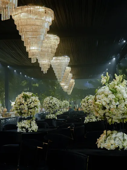 A dramatic, all-black seating arrangement for a modern cocktail event. The space is punctuated by large white floral centerpieces and magnificent tiered crystal chandeliers.