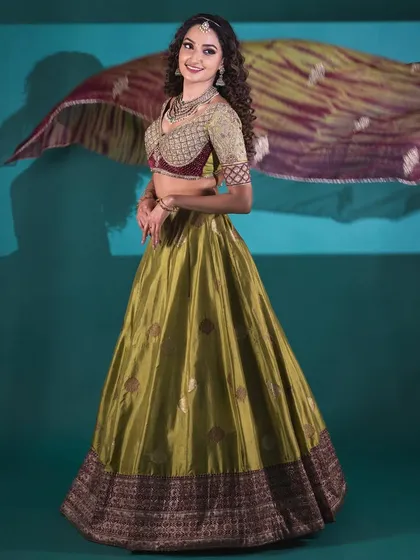 A full view of a custom Banarasi lehenga in a striking olive green. The ensemble features a heavily embroidered blouse with a sweetheart neckline and a flowing skirt, perfect for a sangeet or reception.