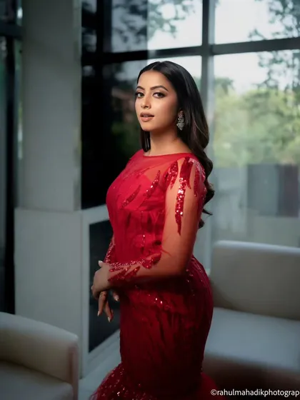 A striking portrait of a woman in a glamorous red gown. The indoor setting with large windows provides soft, natural light that highlights her elegance and the dress's sparkling details.
